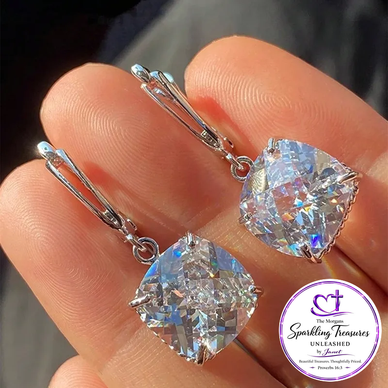 A pair of silver leverback earrings with large iridescent cushion-cut clear gemstones, shown resting on a person's fingers. A circular logo for 'The Morgan Sparkling Treasures Unleashed' is visible in the bottom right corner.