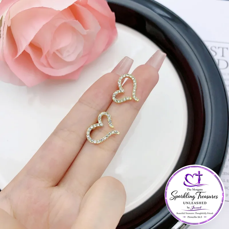 A close-up of a hand wearing two small gold heart-shaped stud earrings with clear rhinestones, set against a white surface with a pink rose and a black dish in the background. A circular logo for 'The Magazine Sparkling Treasures Unleashed by Azzi' is visible in the bottom right corner.