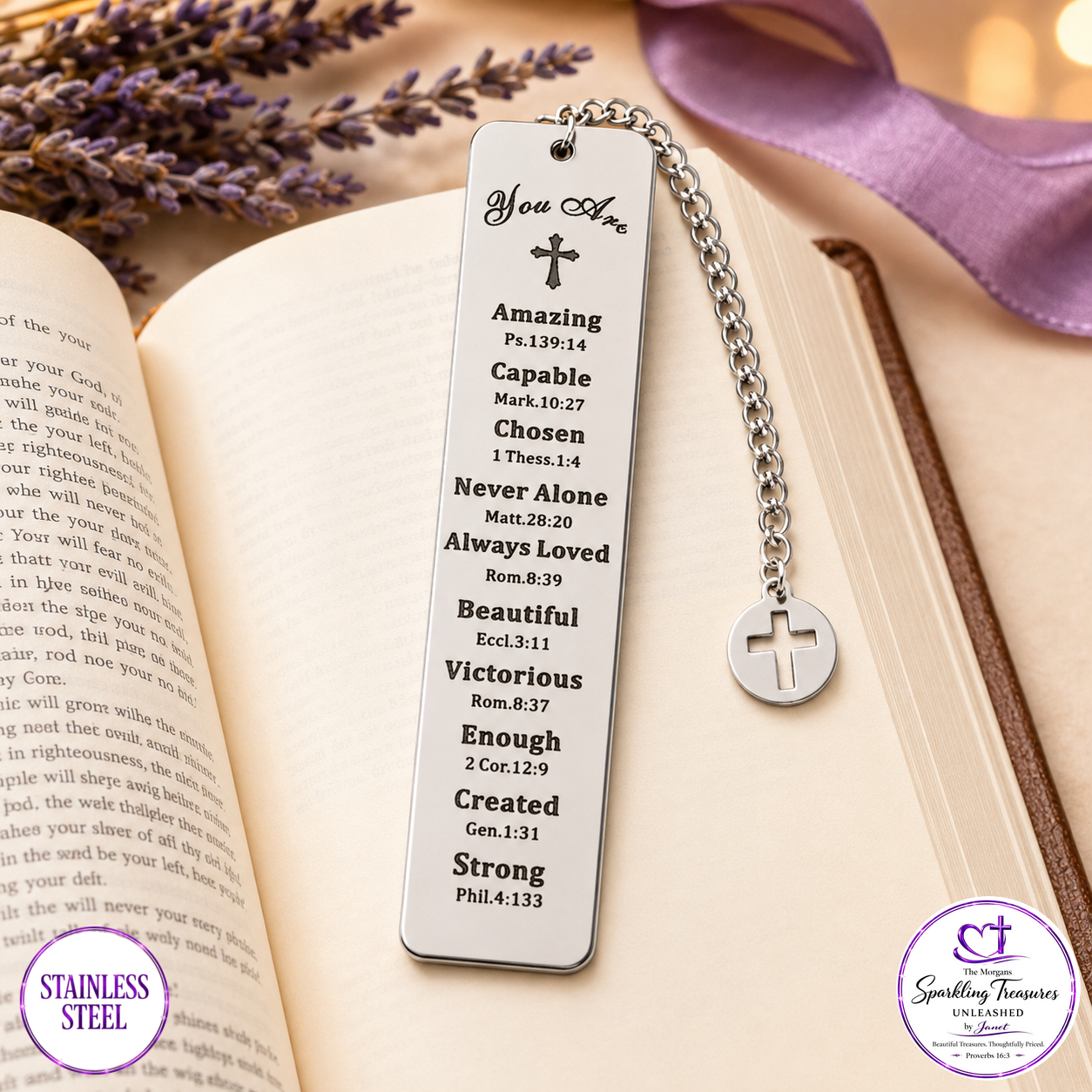 A stainless steel bookmark shaped like a Bible page, engraved with positive affirmations and a small cross at the top, hanging from a silver chain with a round cross pendant. The bookmark is displayed on an open book with lavender and a purple ribbon in the background.