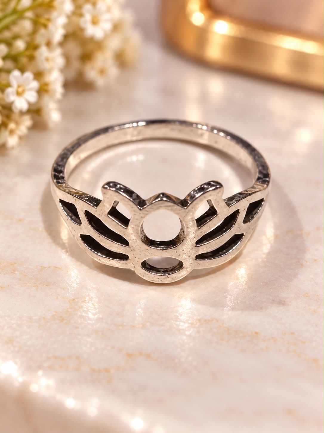 A silver ring with a decorative openwork design, displayed on a light marble surface with small white flowers and a blurred gold object in the background.
