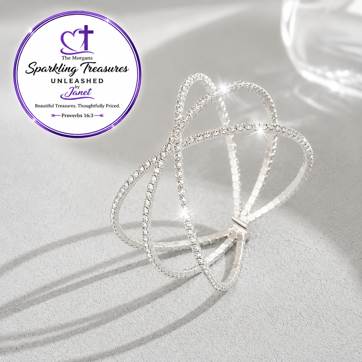 A silver-toned openwork ring with multiple interlocking looped bands, set with small clear stones, photographed on a light gray surface with soft shadows. A purple circular logo in the upper left reads 'The Morgan Sparkling Treasures Unleashed Janet.
