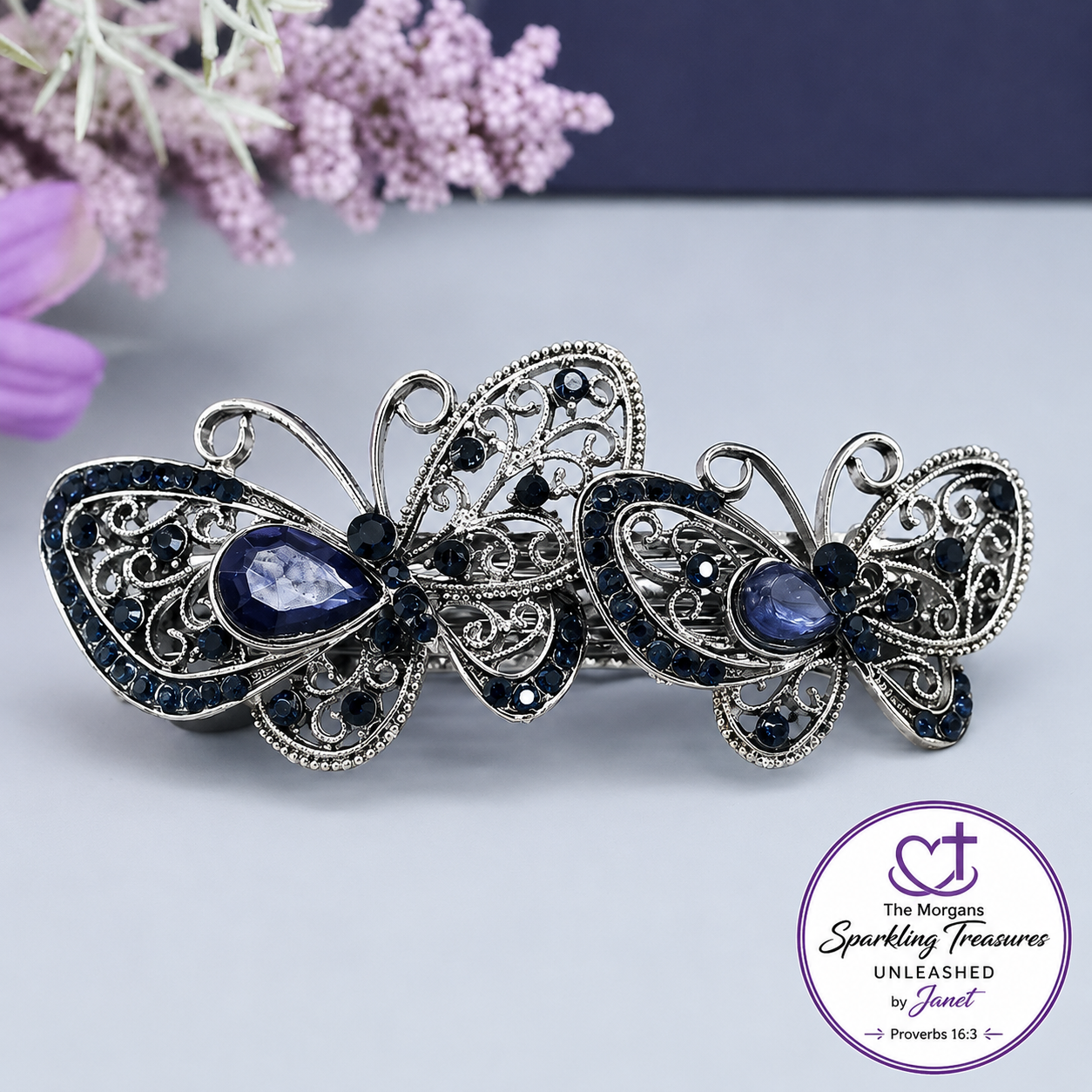 A pair of silver butterfly-shaped earrings with dark blue teardrop gemstones and small dark blue rhinestones, displayed on a light gray surface with a purple floral prop and a circular 'The Morgan Sparkling Treasures Unleashed by Janet' logo in the bottom right corner.