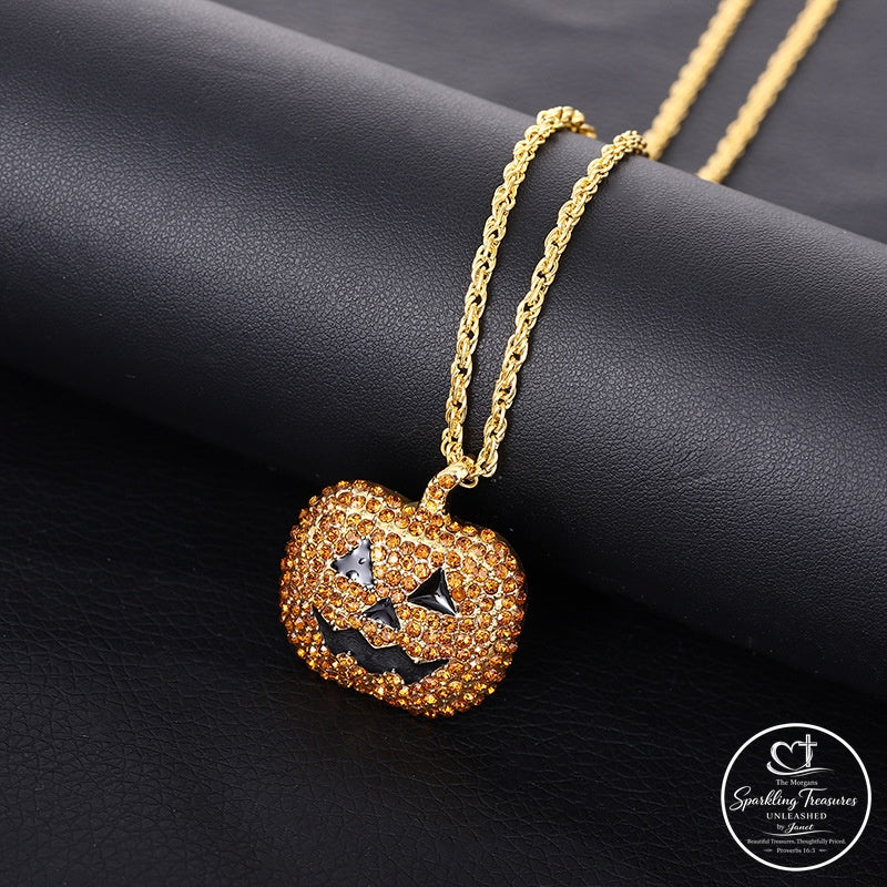 A gold-plated pumpkin pendant necklace covered in small clear rhinestones, with black rhinestone accents forming a jack-o'-lantern face, resting on a black textured surface. A brand watermark for 'Sparkling Treasures' is visible in the bottom right corner.