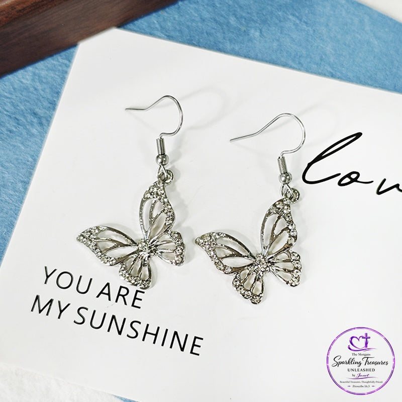 A pair of silver butterfly drop earrings with rhinestone accents, displayed on a white card that reads 'YOU ARE MY SUNSHINE' against a textured blue background.
