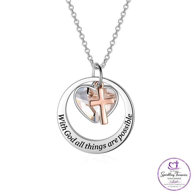 A silver necklace with a circular pendant engraved with 'With God all things are possible' and a rose gold cross inside a clear heart-shaped crystal, shown on a white background with a brand logo in the bottom right corner.