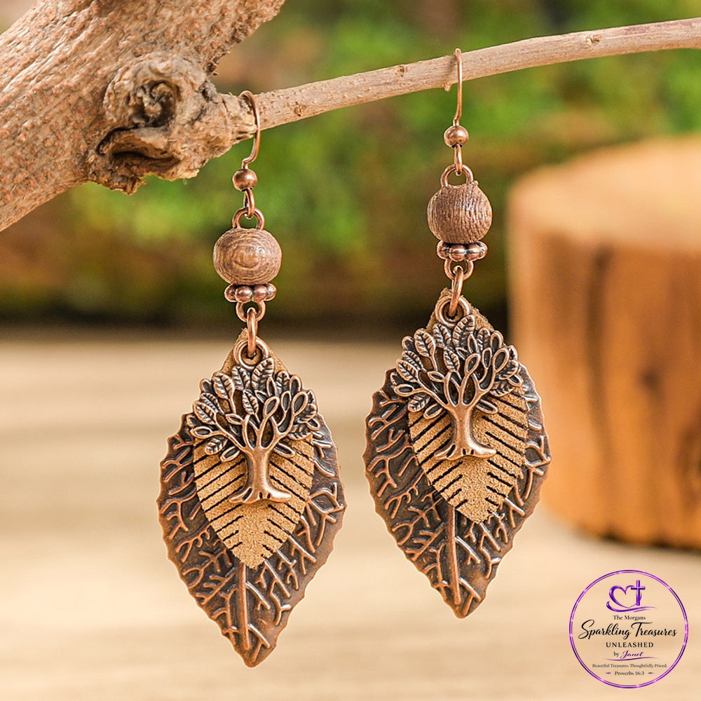 A pair of copper-toned drop earrings shaped like leaves with tree-of-life motifs, hanging from a rustic branch. The background is a softly blurred natural setting with warm wood tones and green foliage.