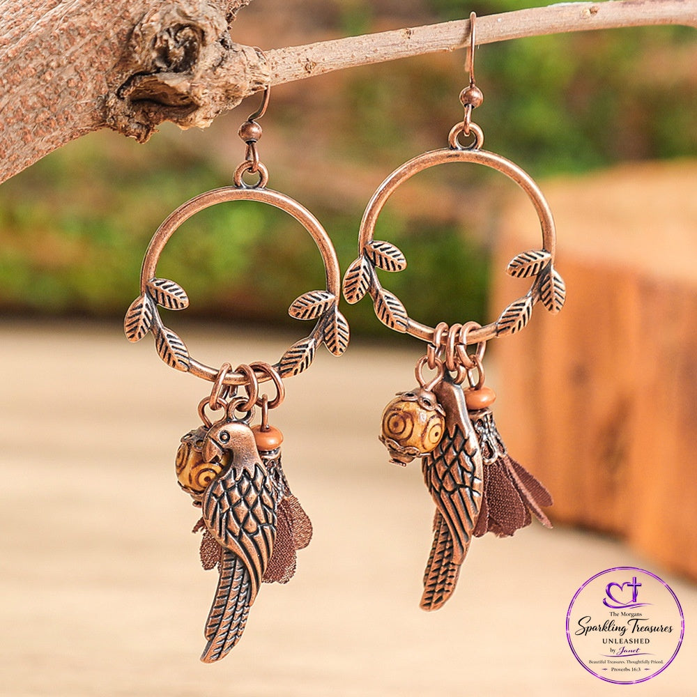 A pair of copper-toned parrot dangle earrings hanging from a rustic tree branch. Each earring features a circular hoop with leafy wire details and a dangling parrot charm with brown feathers and a small gold accent, set against a blurred outdoor background.
