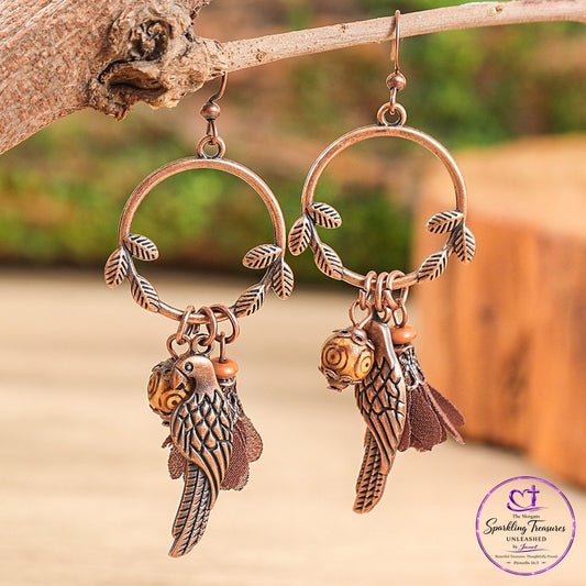 A pair of copper-toned parrot dangle earrings hanging from a rustic tree branch. Each earring features a circular hoop with leafy wire details and a dangling parrot charm with brown feathers and a small gold accent, set against a blurred outdoor background.