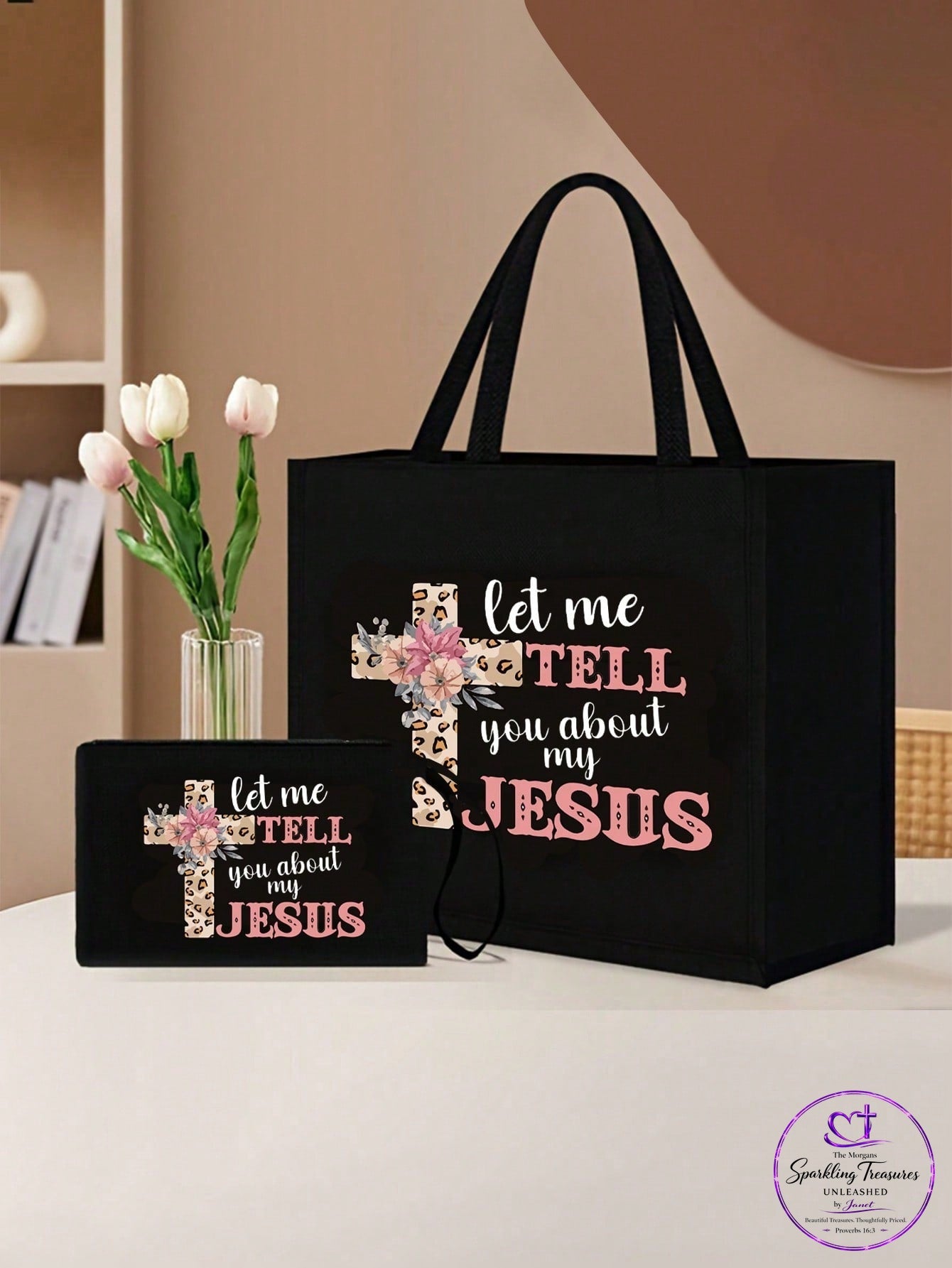 A black tote bag and matching small pouch set with a floral cross graphic and the text 'let me TELL you about my JESUS' on a tabletop. Pink tulips in a vase and a neutral indoor background are visible beside the items.