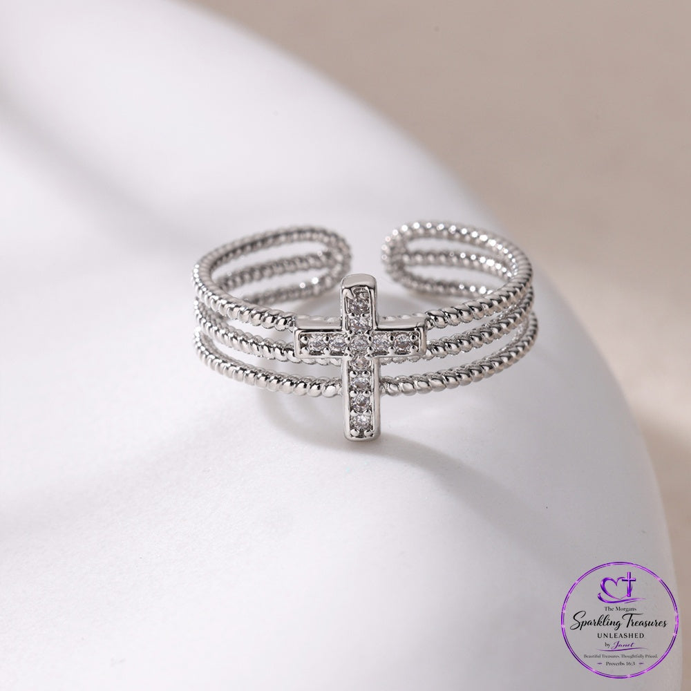 A silver-toned multi-band ring with a central cross charm paved with clear stones, displayed on a white curved surface. The brand logo 'Sparkling Treasures by Laura' is visible in the bottom right corner.