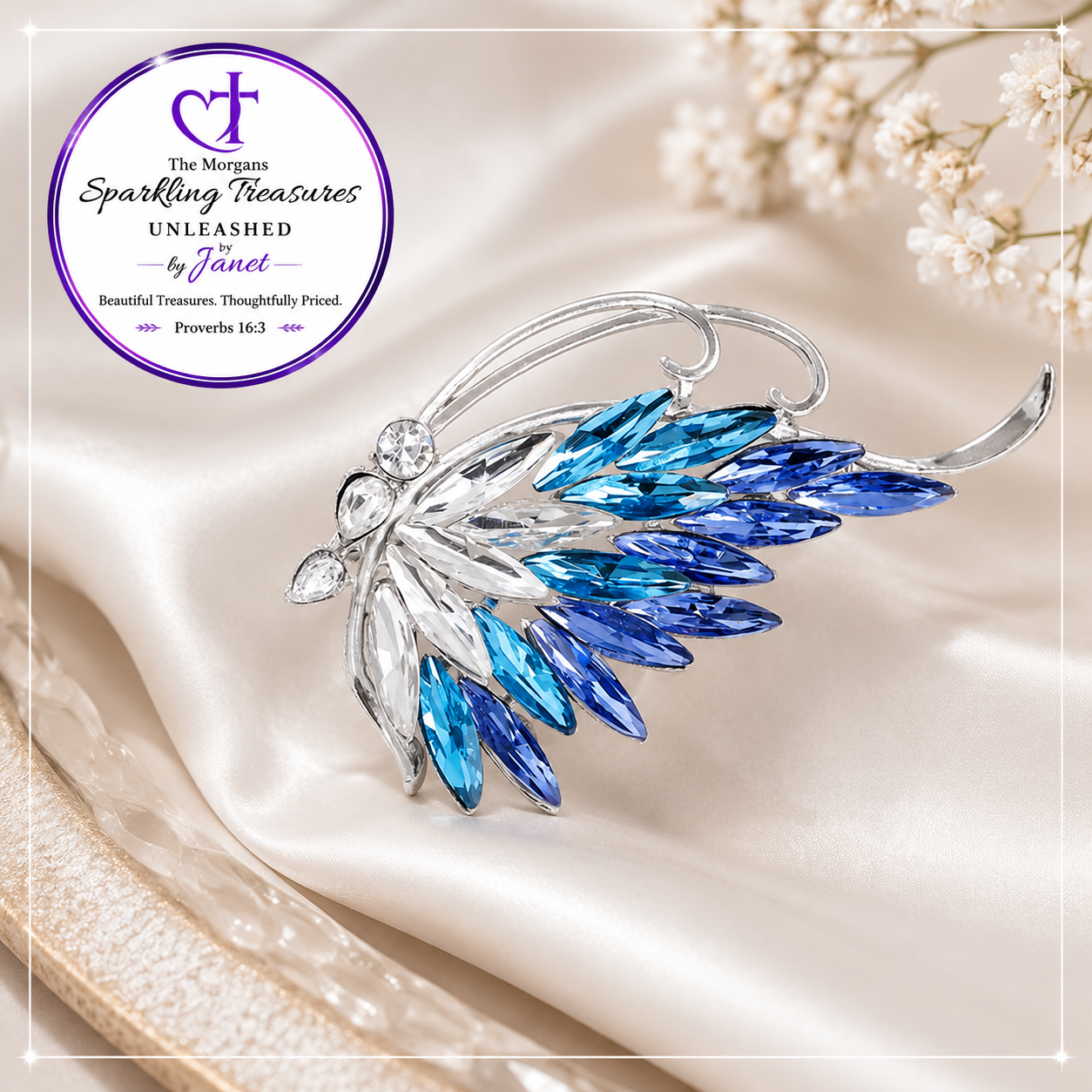 A silver-toned wing-shaped brooch with faceted blue and light blue crystals, displayed on draped cream satin fabric with small white flowers in the corner. A circular logo for 'The Mortals Sparkling Treasures by Janet' appears in the upper left.