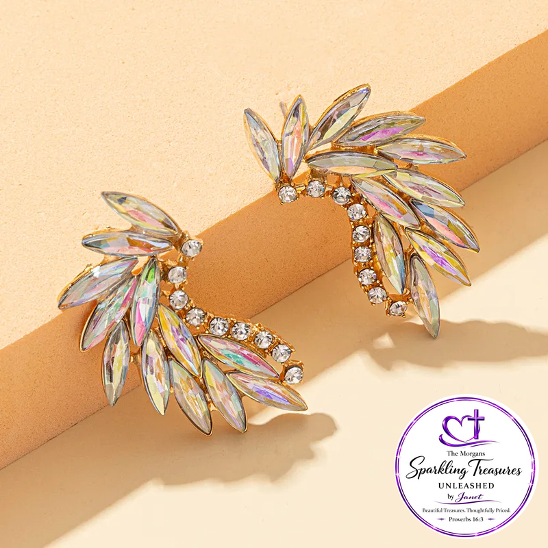 A pair of gold-toned leaf-shaped stud earrings with iridescent marquise crystals and small clear round stones, displayed on a beige surface with a circular 'The Boutique Sparkling Treasures Unleashed by Jewel' logo in the lower right.