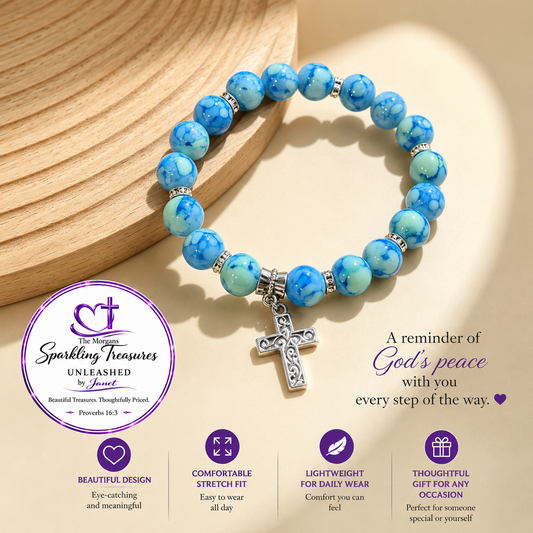 A beaded stretch bracelet with blue marbled round beads, silver spacer beads, and a silver cross charm, displayed on a beige background with a wooden prop. The image includes promotional text and a circular logo for 'The Mother Sparkling Treasures Unleashed by Janet'.