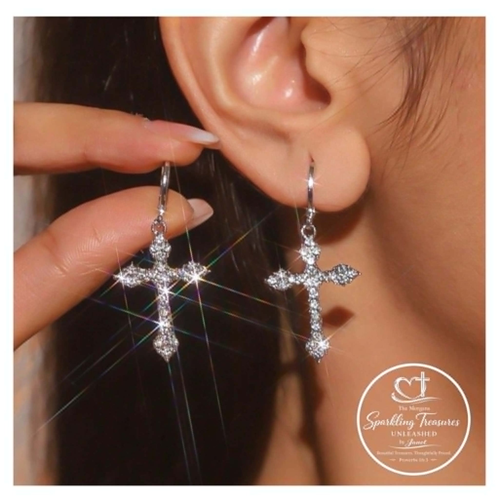 Silver cross earrings being worn and held up to an ear, with a brand logo in the corner.