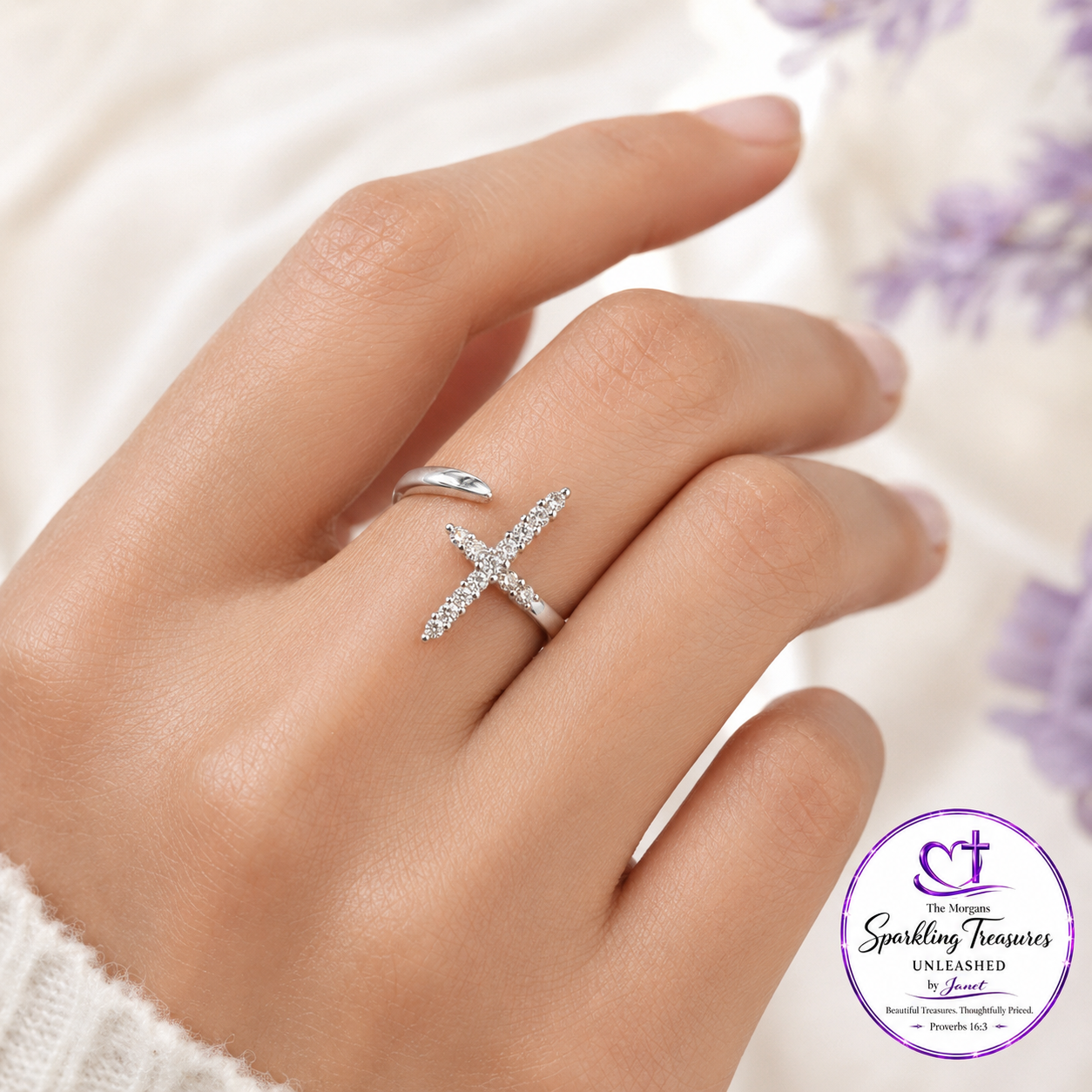 A close-up of a hand wearing a silver cross ring adorned with small clear stones, set against a soft white background with a purple floral corner and a visible Sparkling Treasures logo in the lower right.