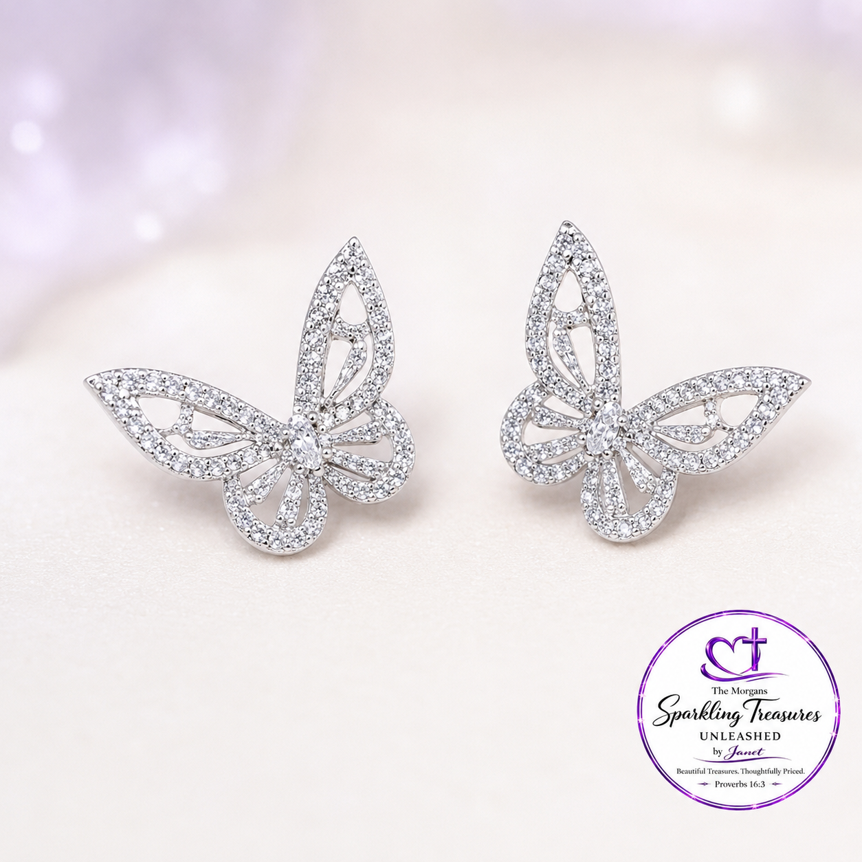 A pair of silver butterfly stud earrings paved with clear crystals, displayed on a soft, light pink textured surface with a brand logo watermark in the bottom right corner.