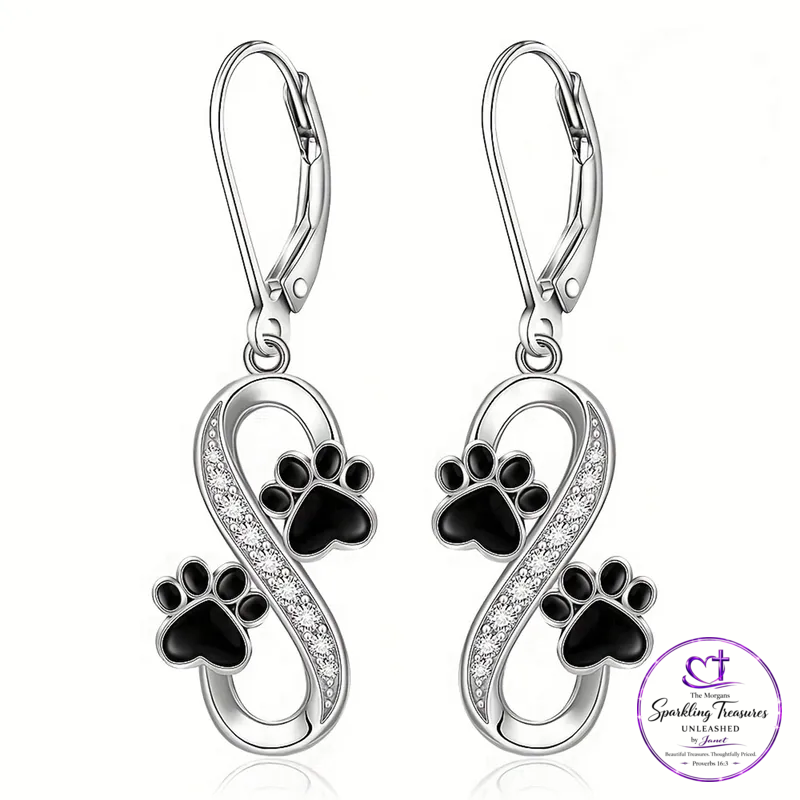 A pair of silver leverback earrings featuring black enamel paw prints and an infinity symbol paved with small clear crystals, shown on a white background with a Sparkling Treasures Unleashed logo in the bottom right corner.