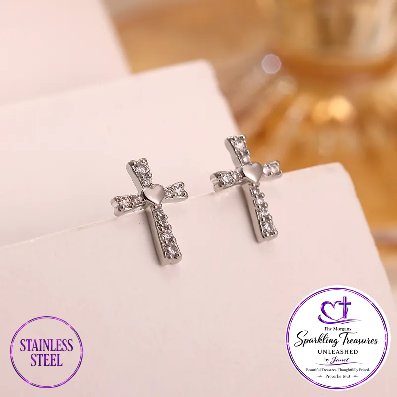 A pair of stainless steel cross stud earrings adorned with clear rhinestones and a small heart in the center, displayed on a white card. The image includes two circular brand badges at the bottom: 'STAINLESS STEEL' on the left and 'The Morgan Sparkling Treasures UNLEASHED by Anusha' on the right.