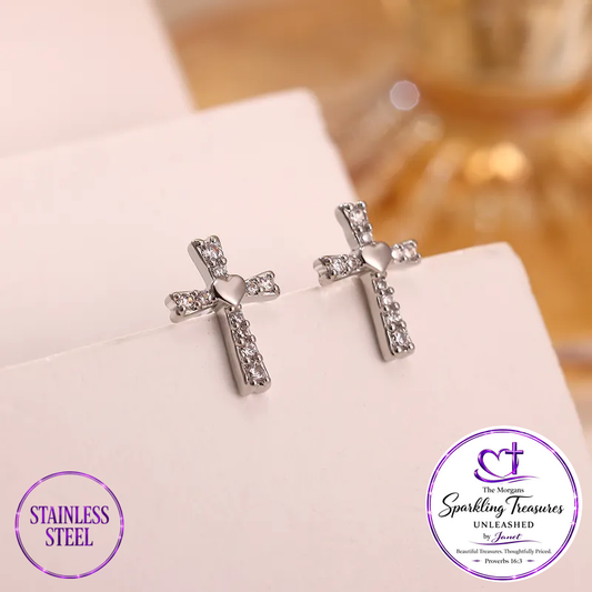 A pair of stainless steel cross stud earrings adorned with clear rhinestones and a small heart in the center, displayed on a white card. The image includes two circular brand badges at the bottom: 'STAINLESS STEEL' on the left and 'The Morgan Sparkling Treasures UNLEASHED by Anusha' on the right.