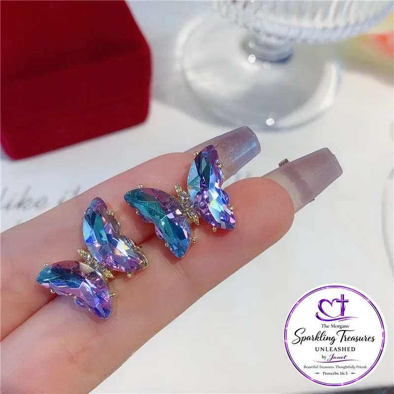 Three butterfly-shaped rhinestone nail art stickers displayed on a person's fingernails, with a red jewelry box and glass base in the background. A 'The Mignon Sparkling Treasures Unleashed' logo is visible in the bottom right corner.