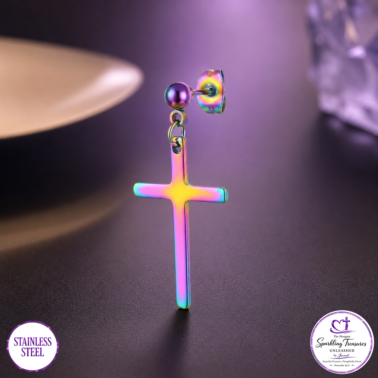 A single drop earring featuring a stainless steel cross pendant with an iridescent rainbow finish, hanging from a round stud. The background is a dark, moody surface with purple lighting, and two circular brand badges are visible in the bottom corners.