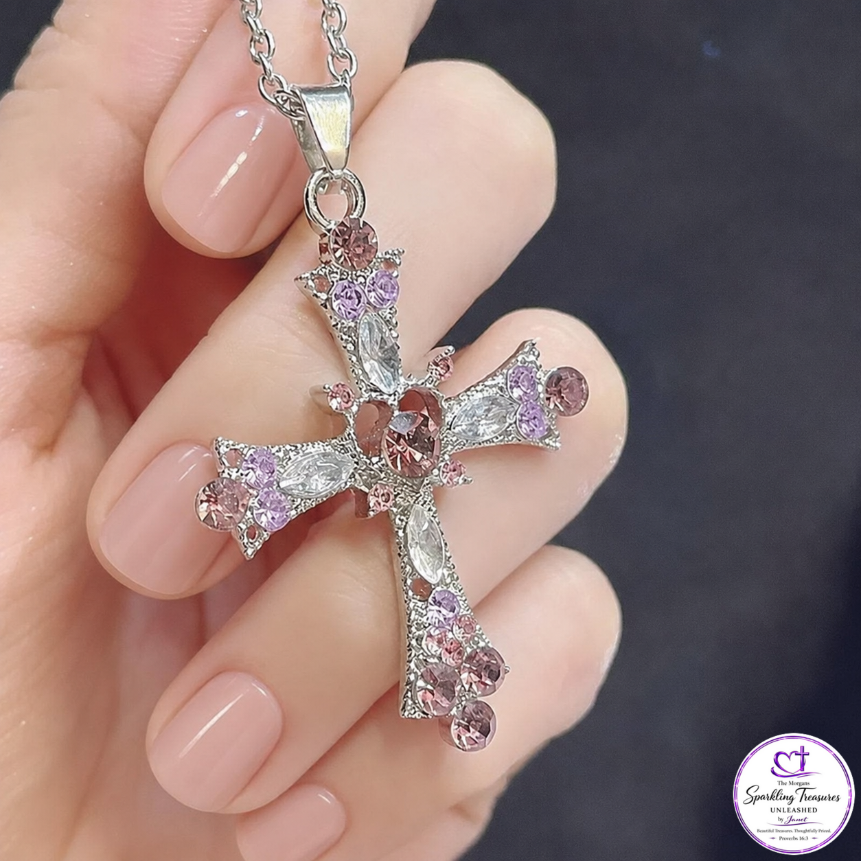 A silver-toned cross pendant necklace adorned with clear marquise crystals and pink round crystals, held in a hand with manicured nails against a dark background.
